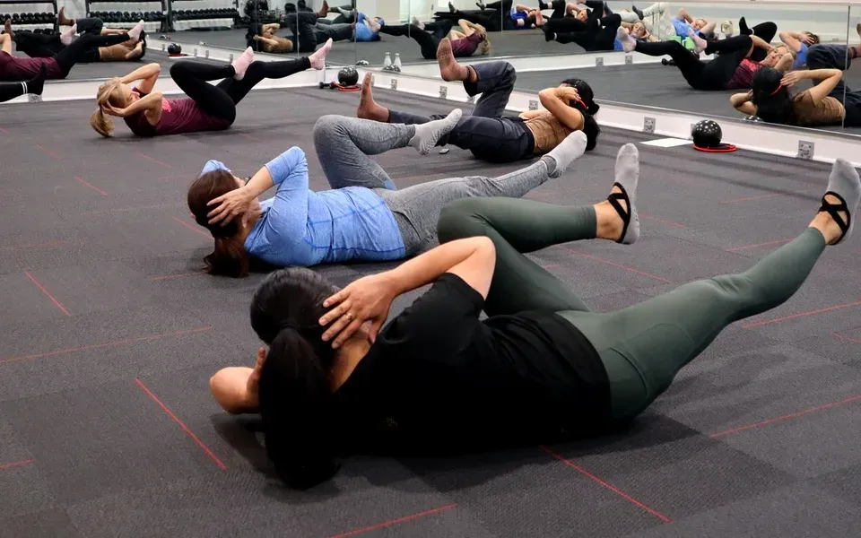 Un groupe fait des exercices d’abdominaux allongé sur un sol gris dans une salle à miroirs