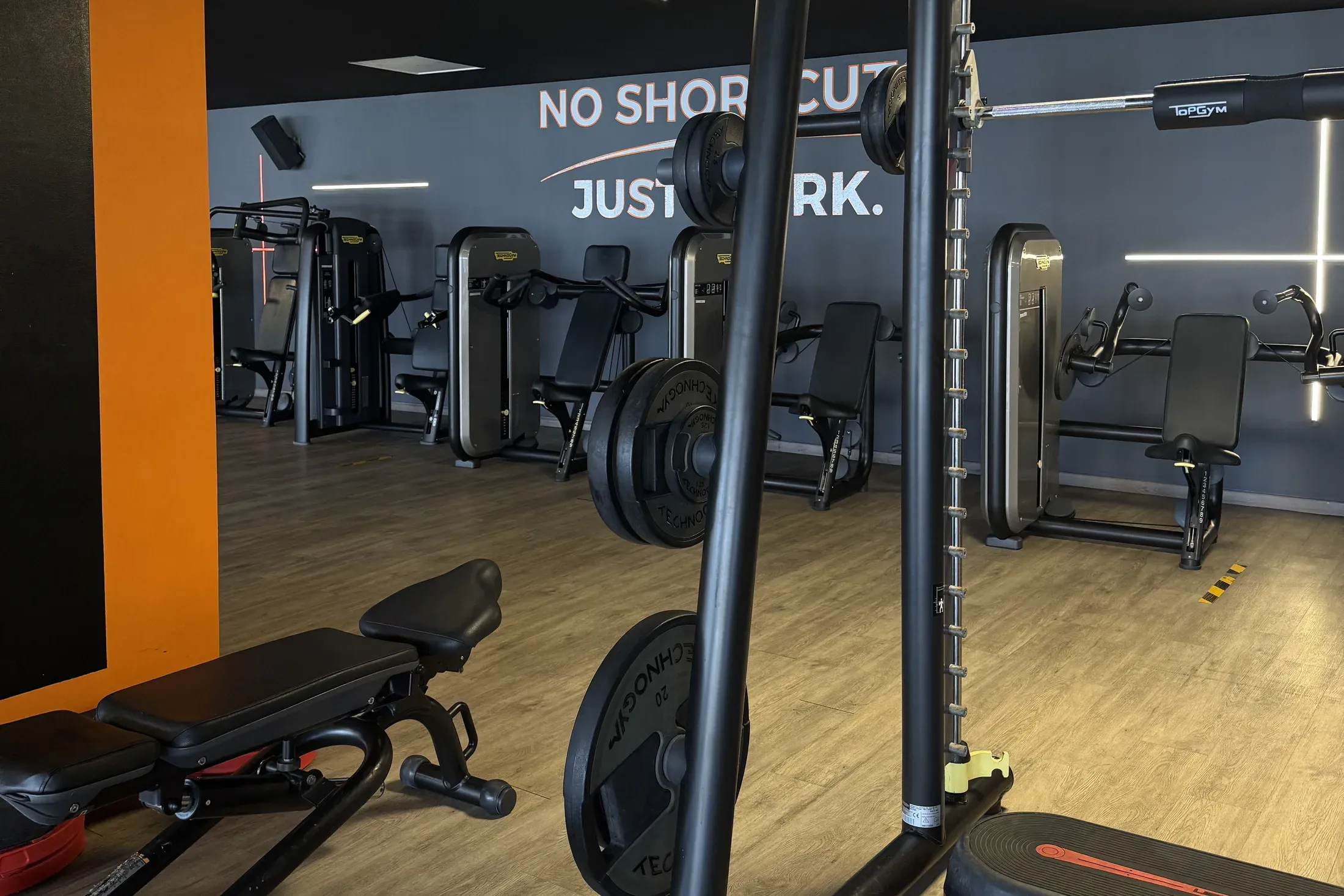 Salle de musculation : bancs réglables, cage à squat et machines devant le mur « No shortcuts, just work »